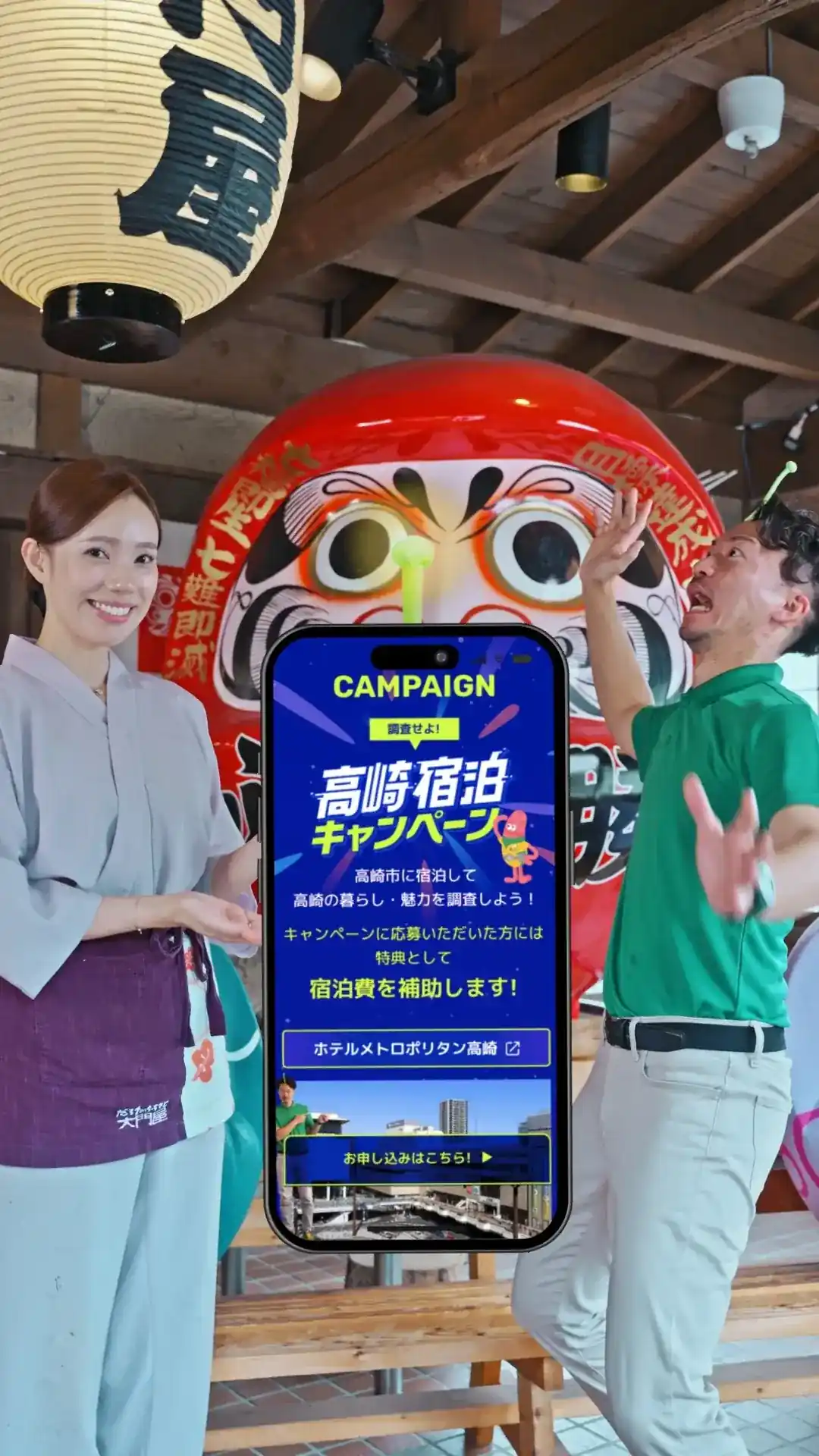 高崎宿泊キャンペーンの告知画面を持つスタッフ(だるまの前で)